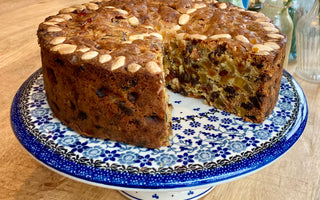 Kersttaart op een Bunzlau Castle cakestand. Er is een stuk uit de taart gesneden. Je ziet de bovenkant en een deel van de binnenkant. De taart is bruin met bovenop amandelen en aan de binnenkant gekonfijte vruchtjes.