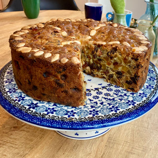 Kersttaart op een Bunzlau Castle cakestand. Er is een stuk uit de taart gesneden. Je ziet de bovenkant en een deel van de binnenkant. De taart is bruin met bovenop amandelen en aan de binnenkant gekonfijte vruchtjes.