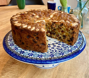 Kersttaart op een Bunzlau Castle cakestand. Er is een stuk uit de taart gesneden. Je ziet de bovenkant en een deel van de binnenkant. De taart is bruin met bovenop amandelen en aan de binnenkant gekonfijte vruchtjes.