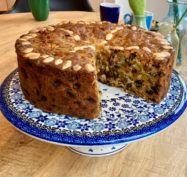 Kersttaart op een Bunzlau Castle cakestand. Er is een stuk uit de taart gesneden. Je ziet de bovenkant en een deel van de binnenkant. De taart is bruin met bovenop amandelen en aan de binnenkant gekonfijte vruchtjes.