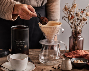 Met een schepje wordt gemalen koffie in een filter gedaan. Het papieren filter zit in een glazen filterhouder, op een glazen koffiekan. Op tafel staan verder het koffieblik met gemalen koffie, een kop-en-schotel en een vaas met droogbloemen.